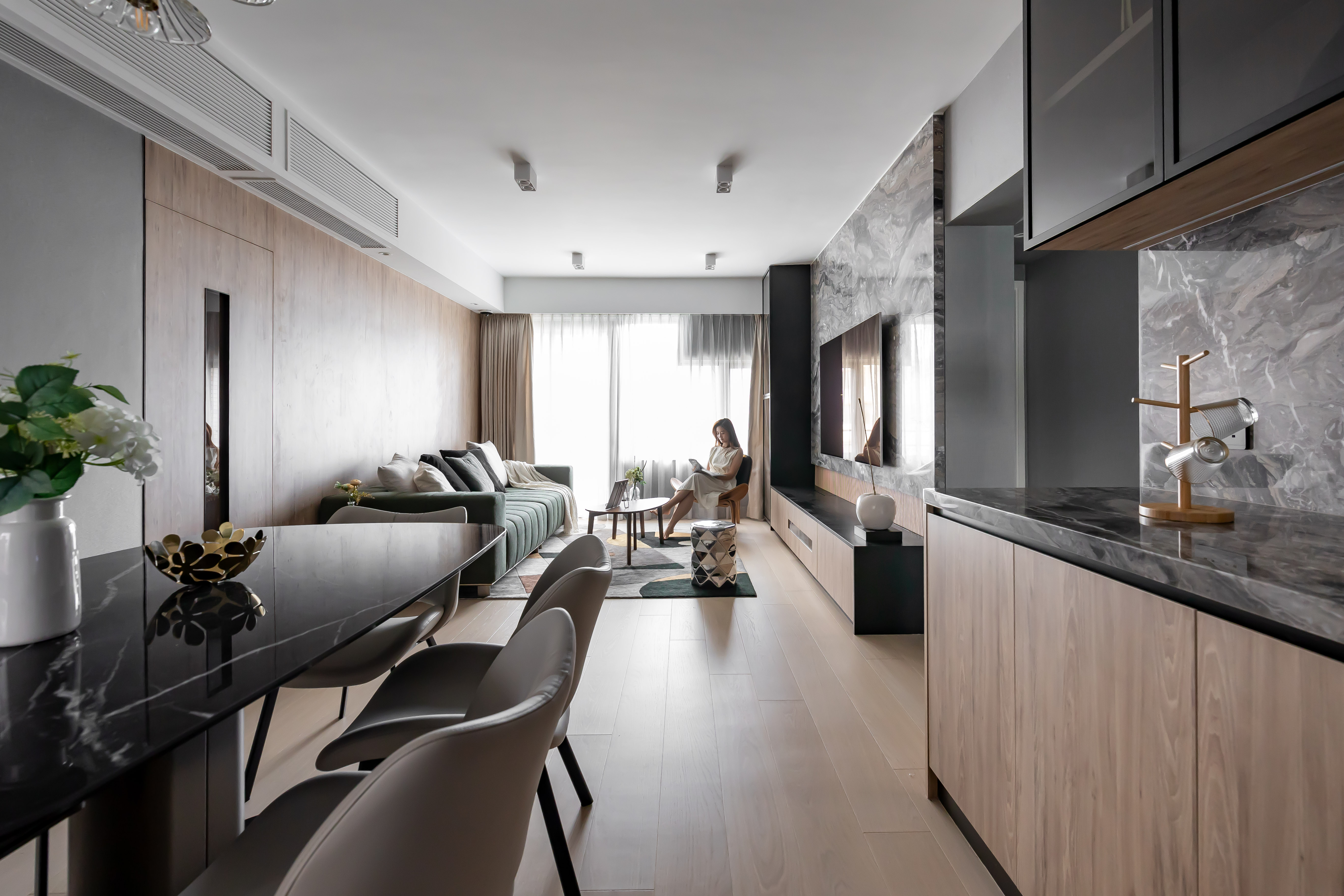 Modern open-plan living and dining room with a black marble dining table, green sofa, and a woman sitting by the window.