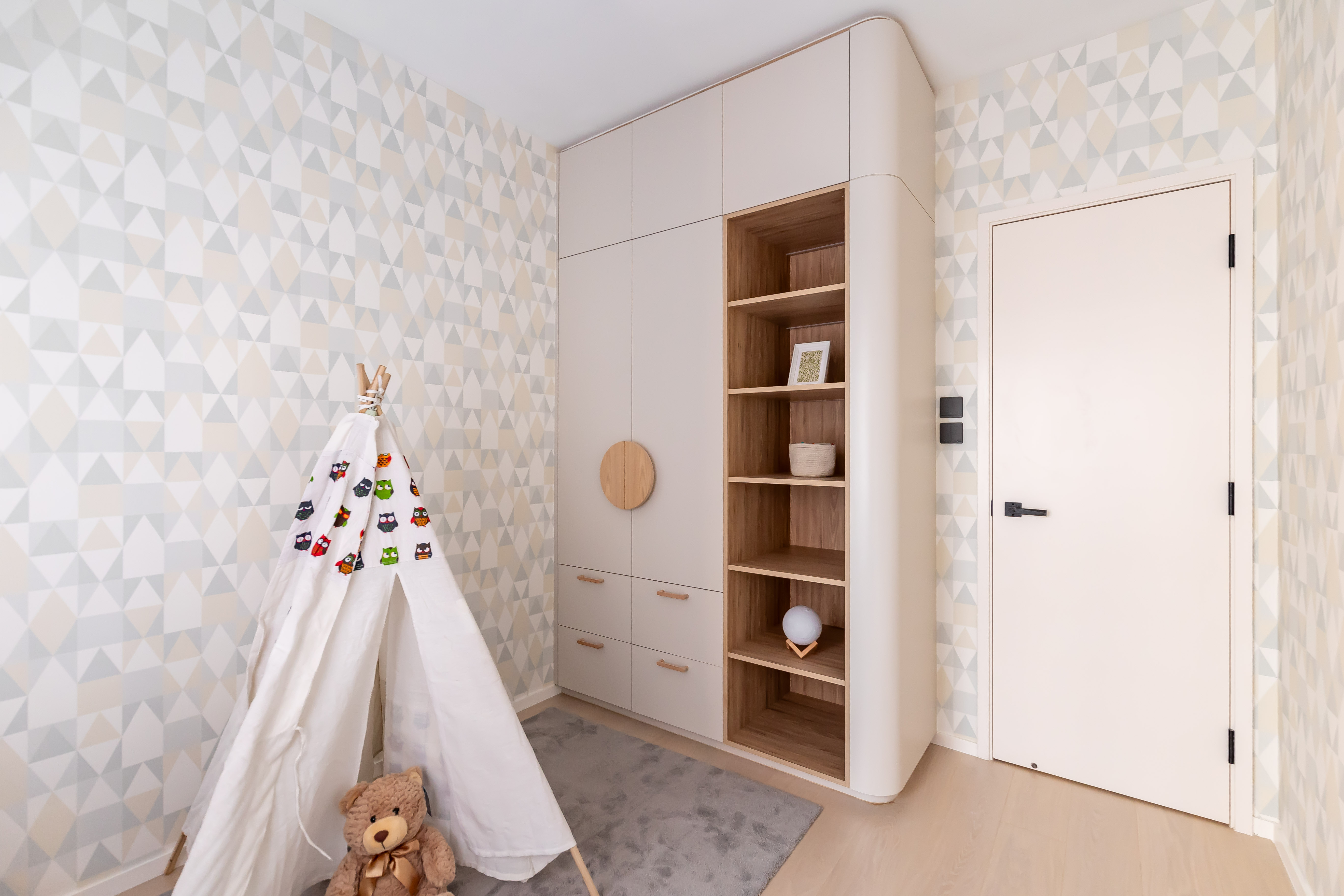 Children's room with geometric wallpaper, a white curved wardrobe with wooden shelves, and a white teepee tent with a teddy bear.