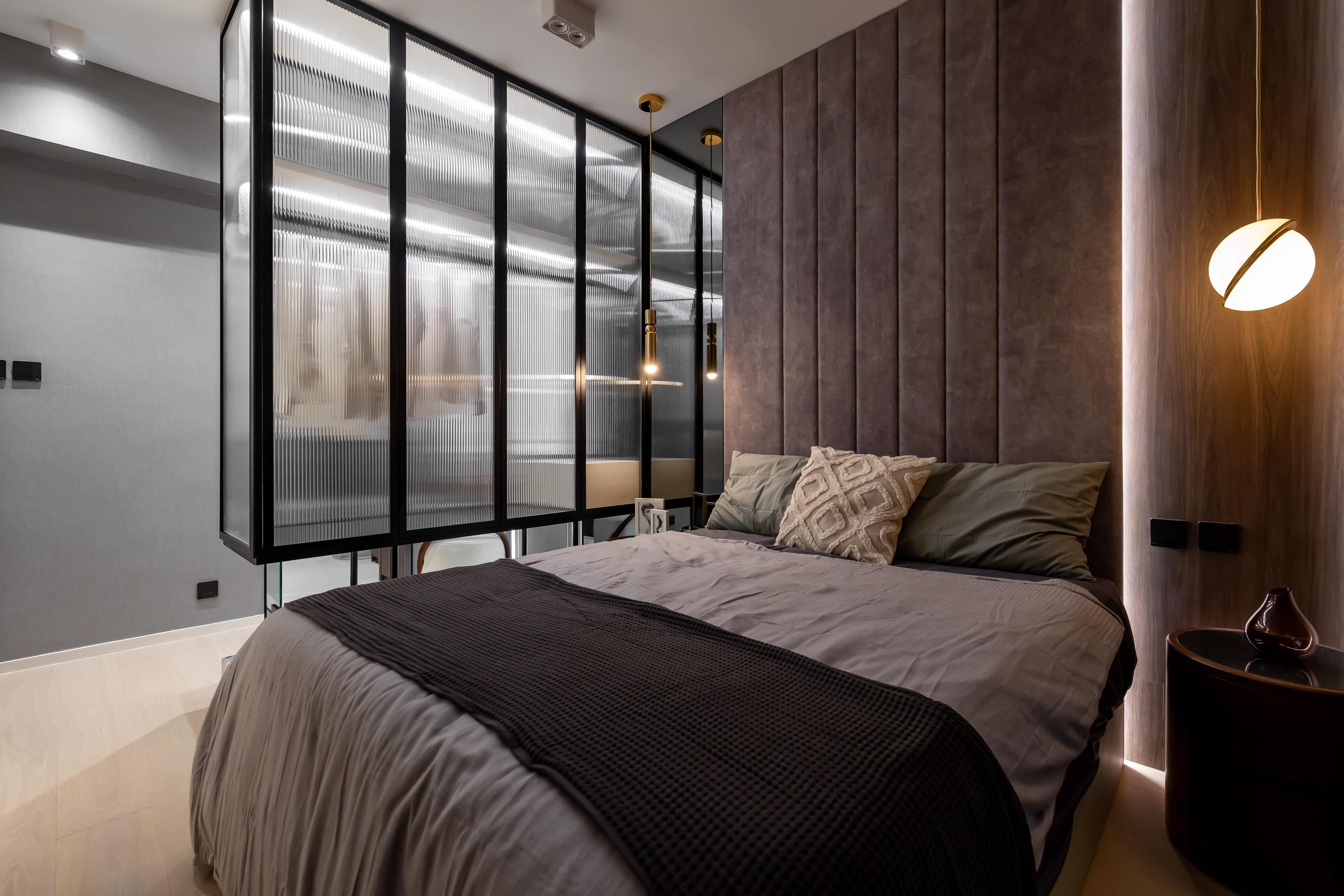 A modern bedroom featuring a bed with grey bedding, a padded headboard, and a fluted glass wardrobe partition.