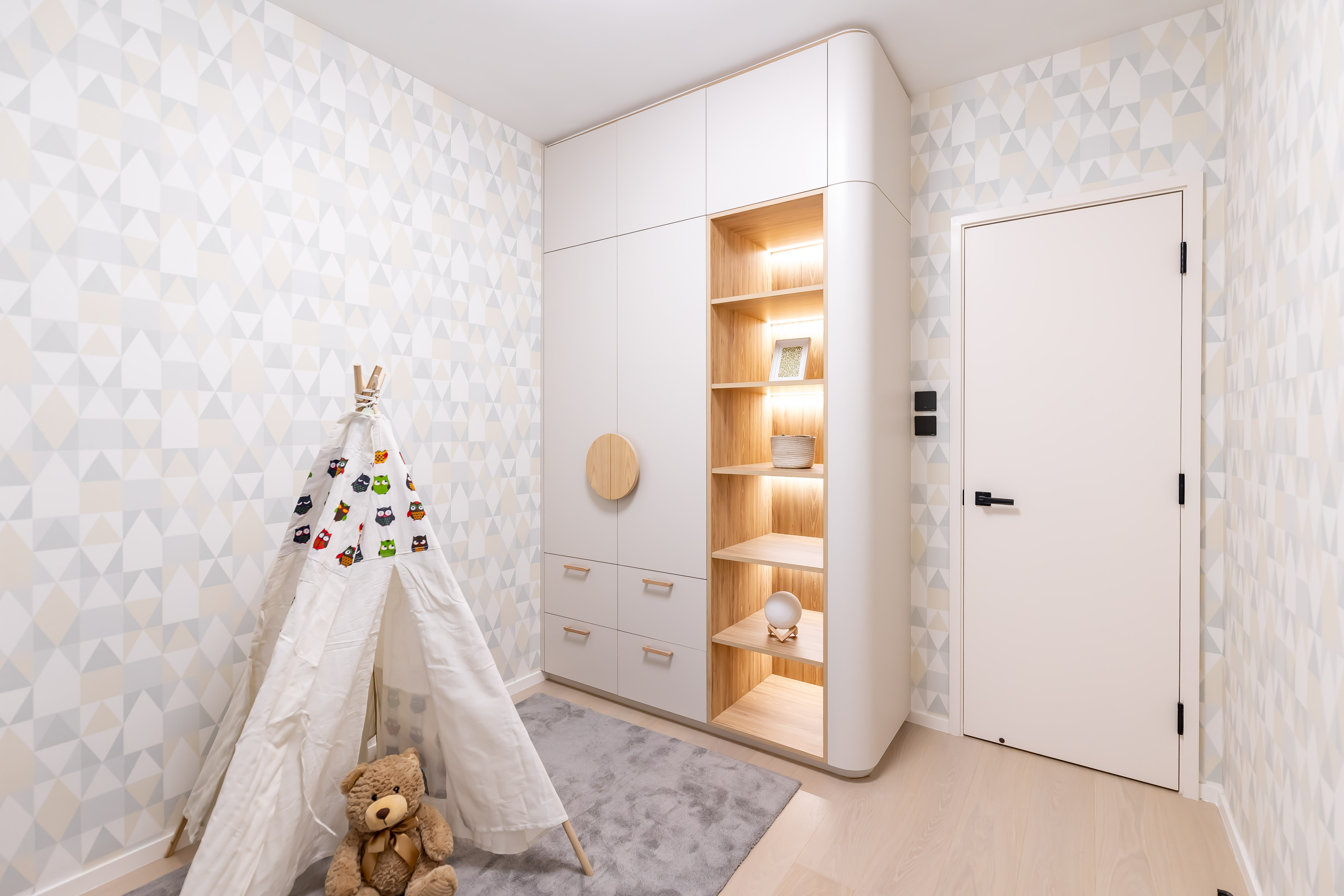 A children's room featuring a white and wood wardrobe, a play tent, a teddy bear, and geometric patterned wallpaper.