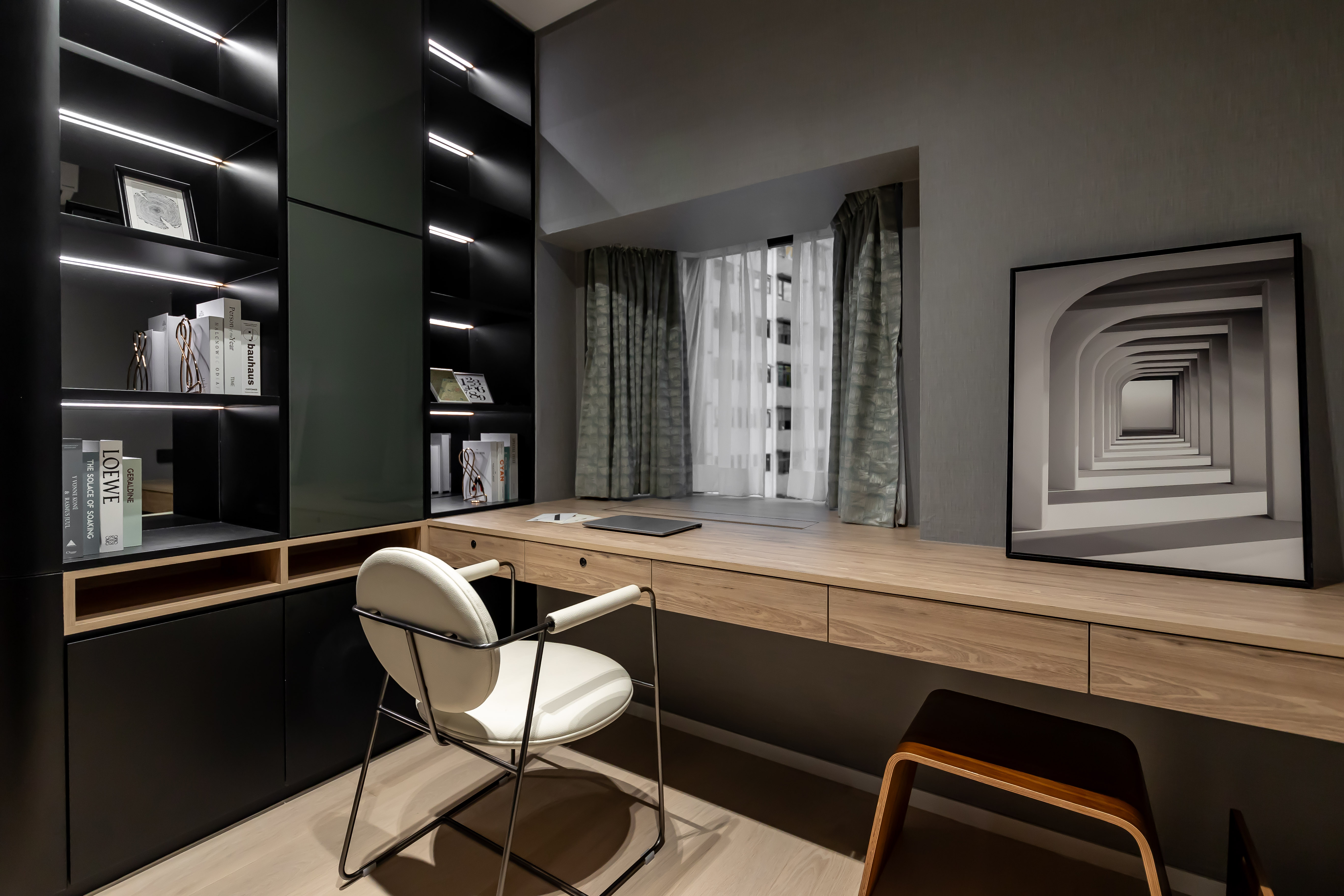 A modern study room featuring a long wooden desk, black display shelves, a white leather chair, and a framed architectural print.