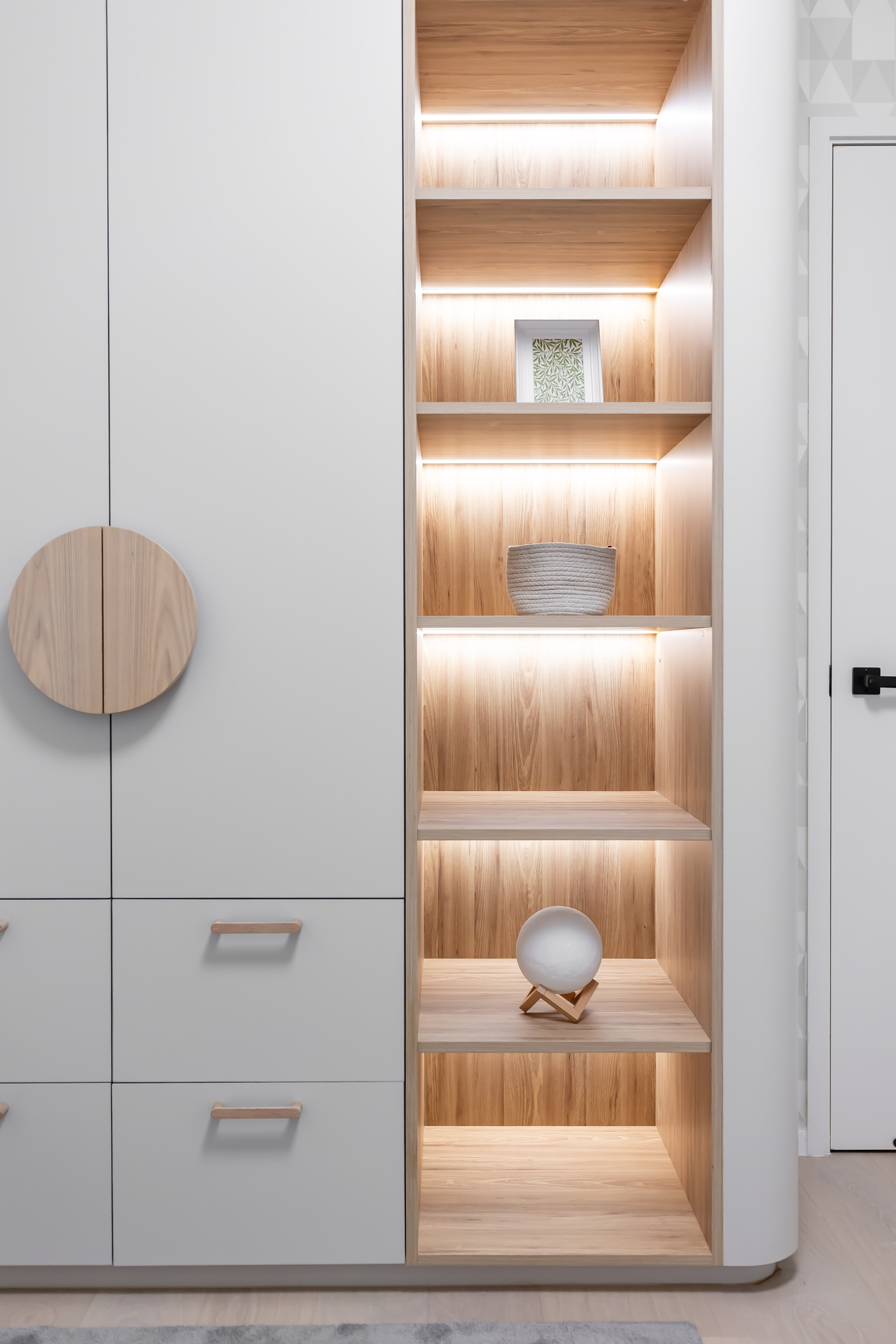 White built-in wardrobe with wooden handles and illuminated open wooden shelving displaying small decorative items.