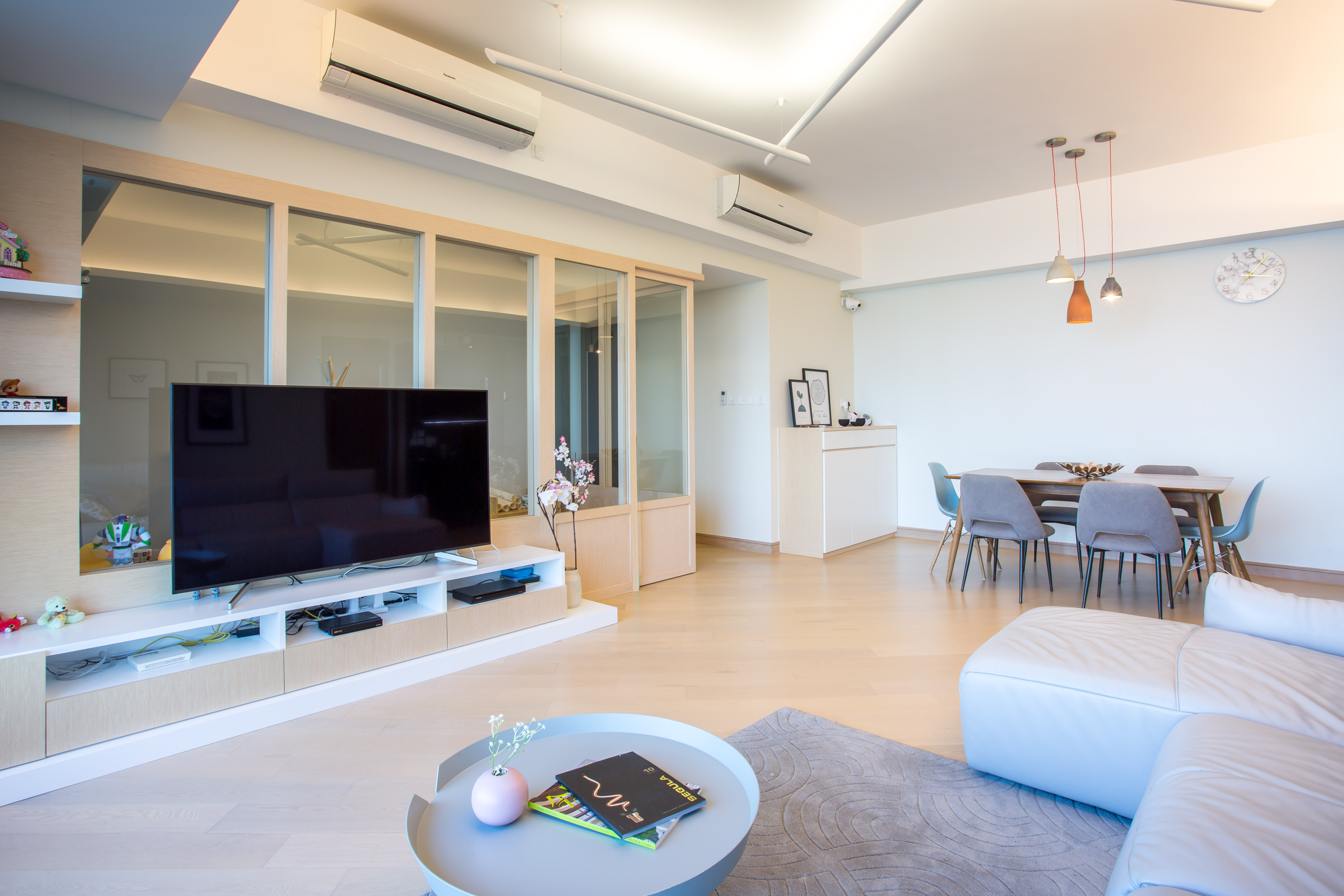 Spacious living and dining room with a grey sofa, TV, glass partition, and a wooden dining table with grey chairs.