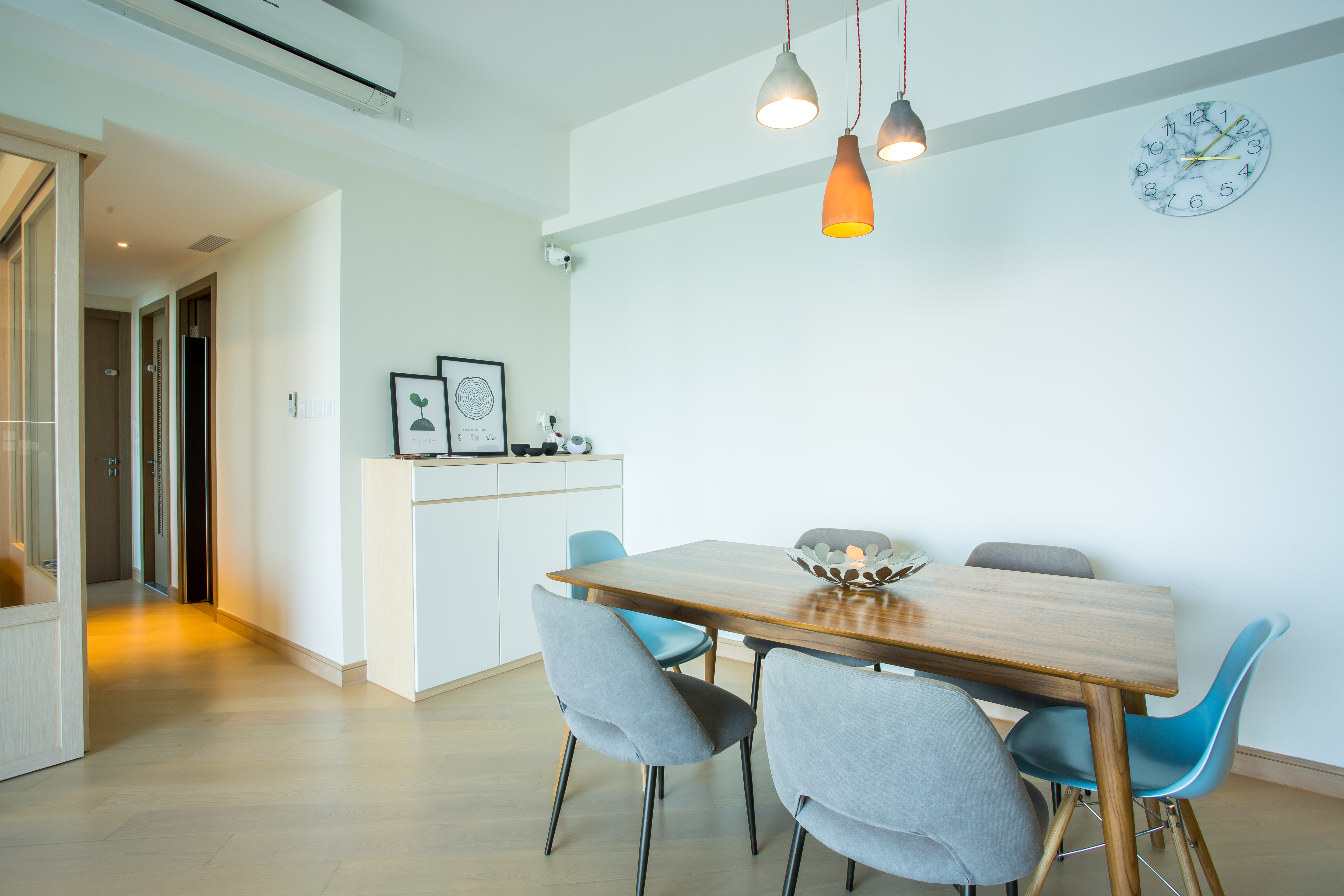 A dining room with a wooden table, grey and blue chairs, three pendant lights, and a white cabinet.