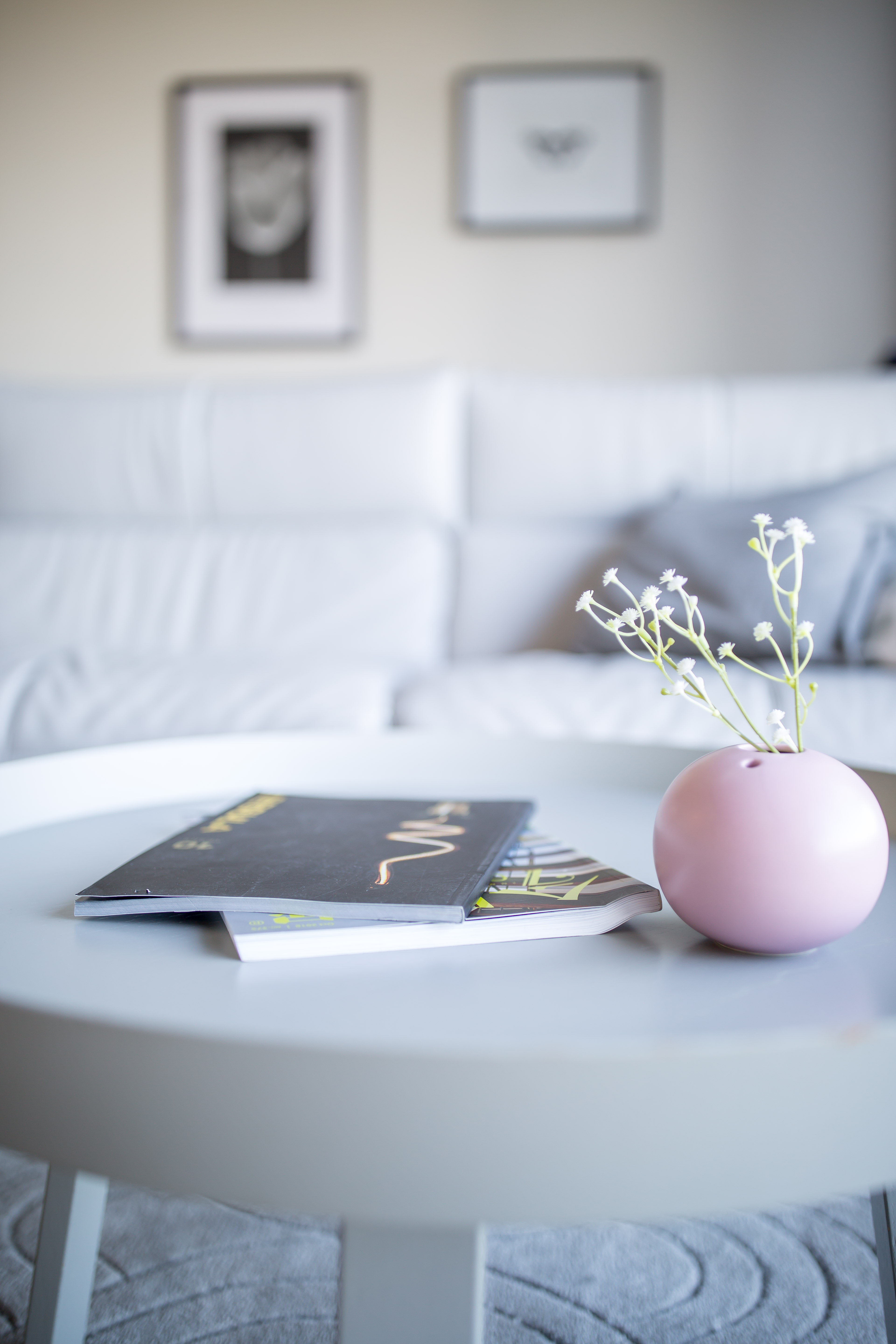 A white coffee table with two magazines and a small pink vase holding white flowers, with a white sofa in the background.