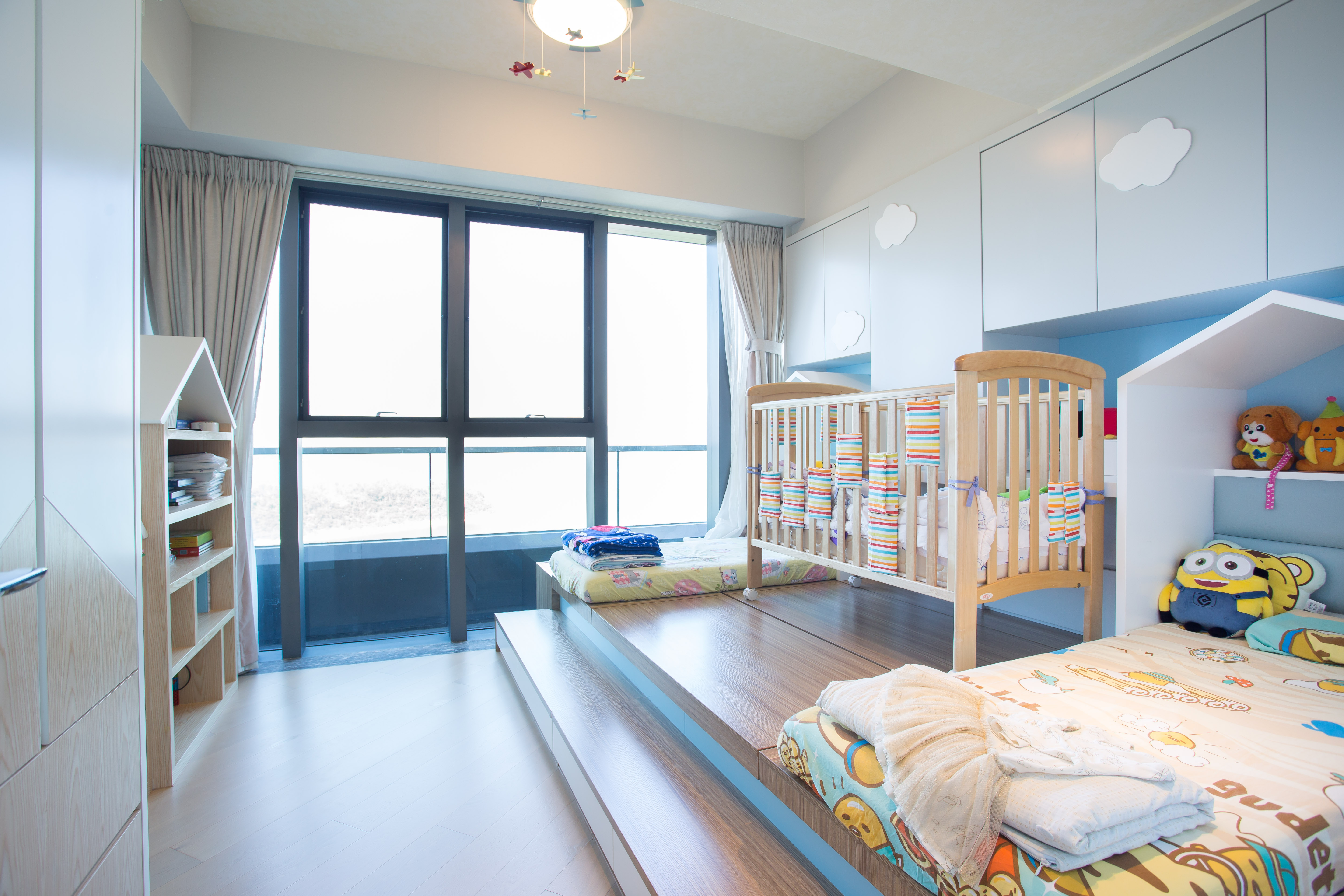 Children's bedroom featuring a wooden platform, baby crib, light blue cloud-decorated cabinets, and a house-shaped bookshelf.