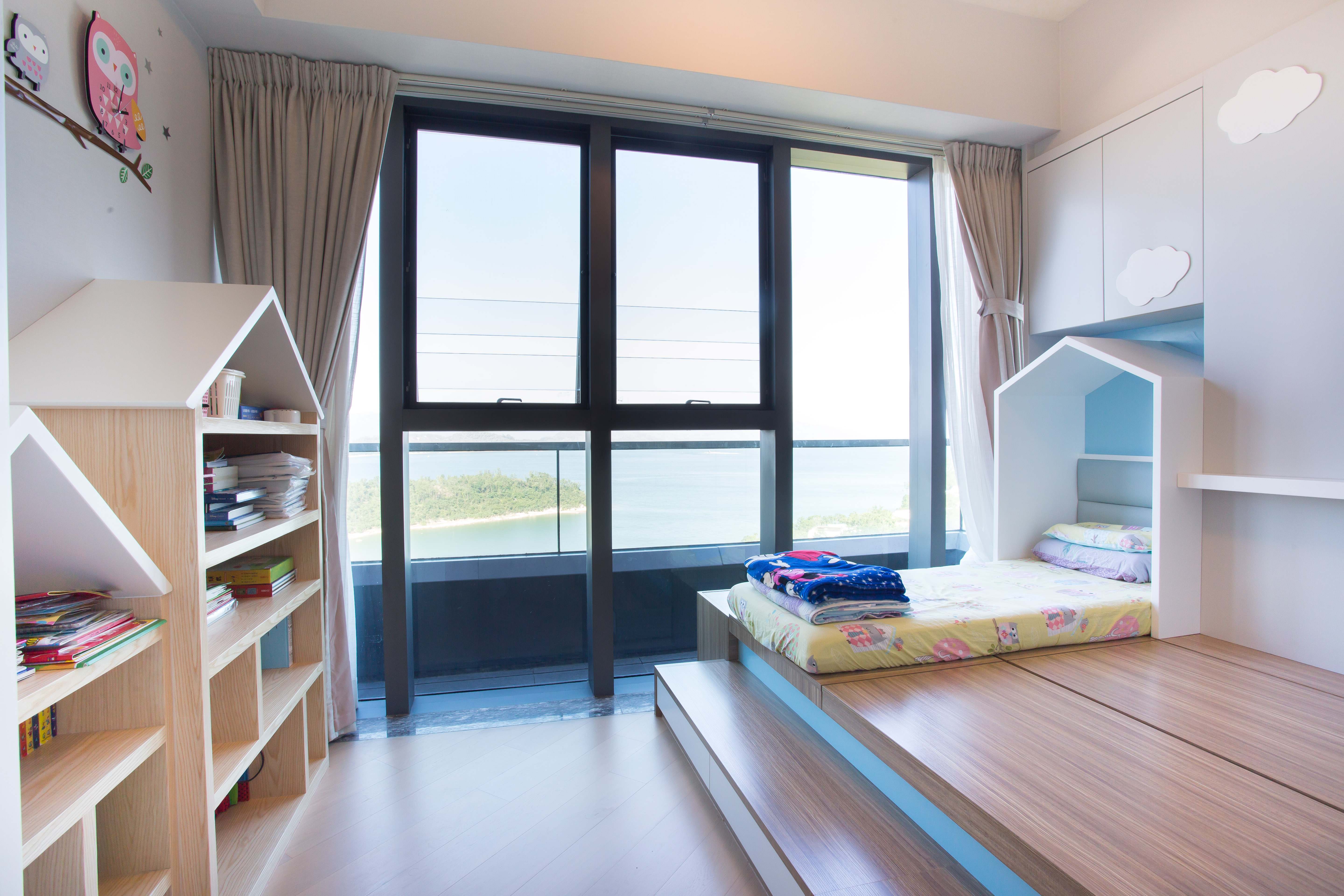 Children's room with a wooden platform bed, house-shaped headboard, house-shaped bookshelf, and a large window showing a sea view.