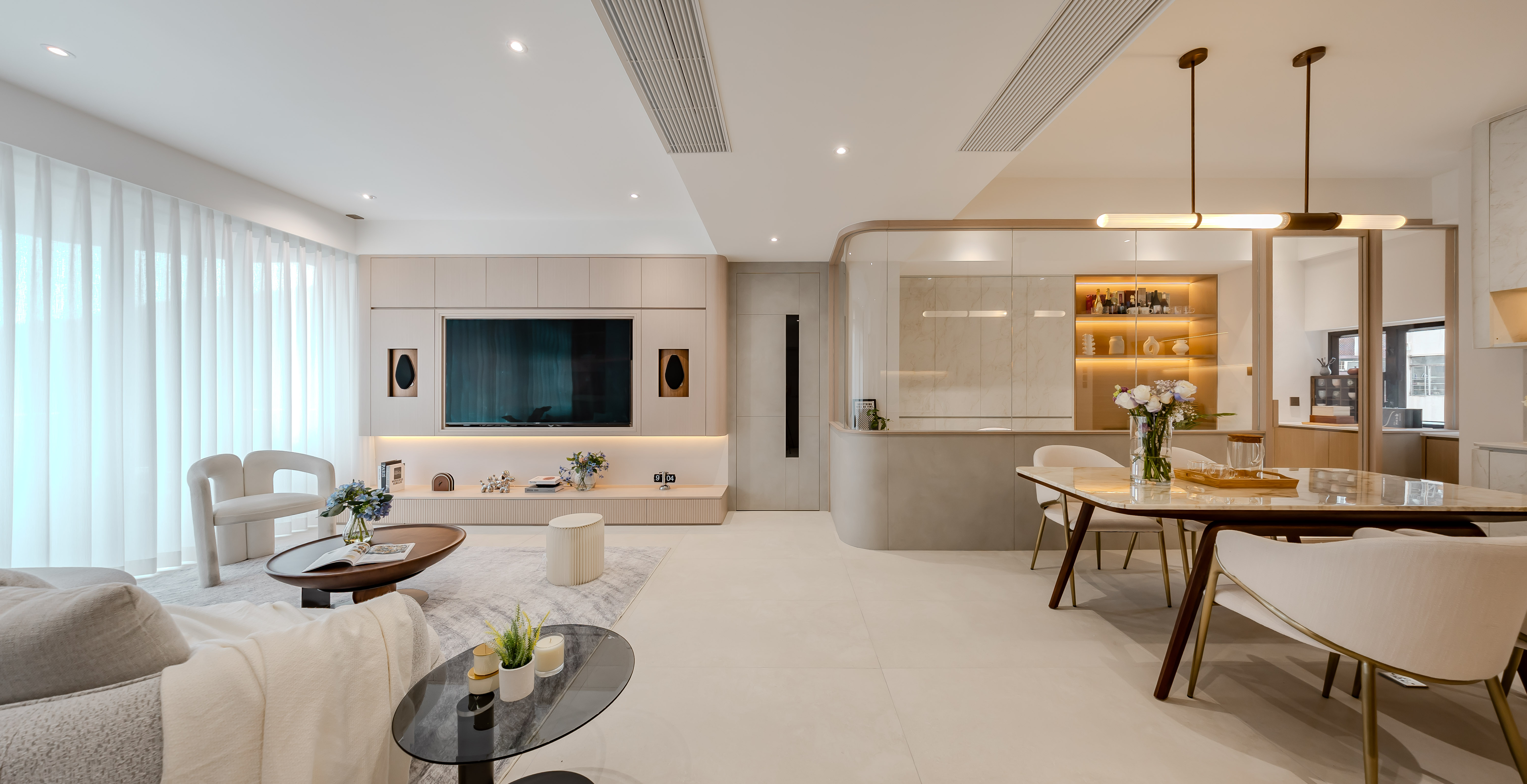 Spacious living and dining room featuring a large TV wall, light-colored sofa, marble dining table, and a glass partition.