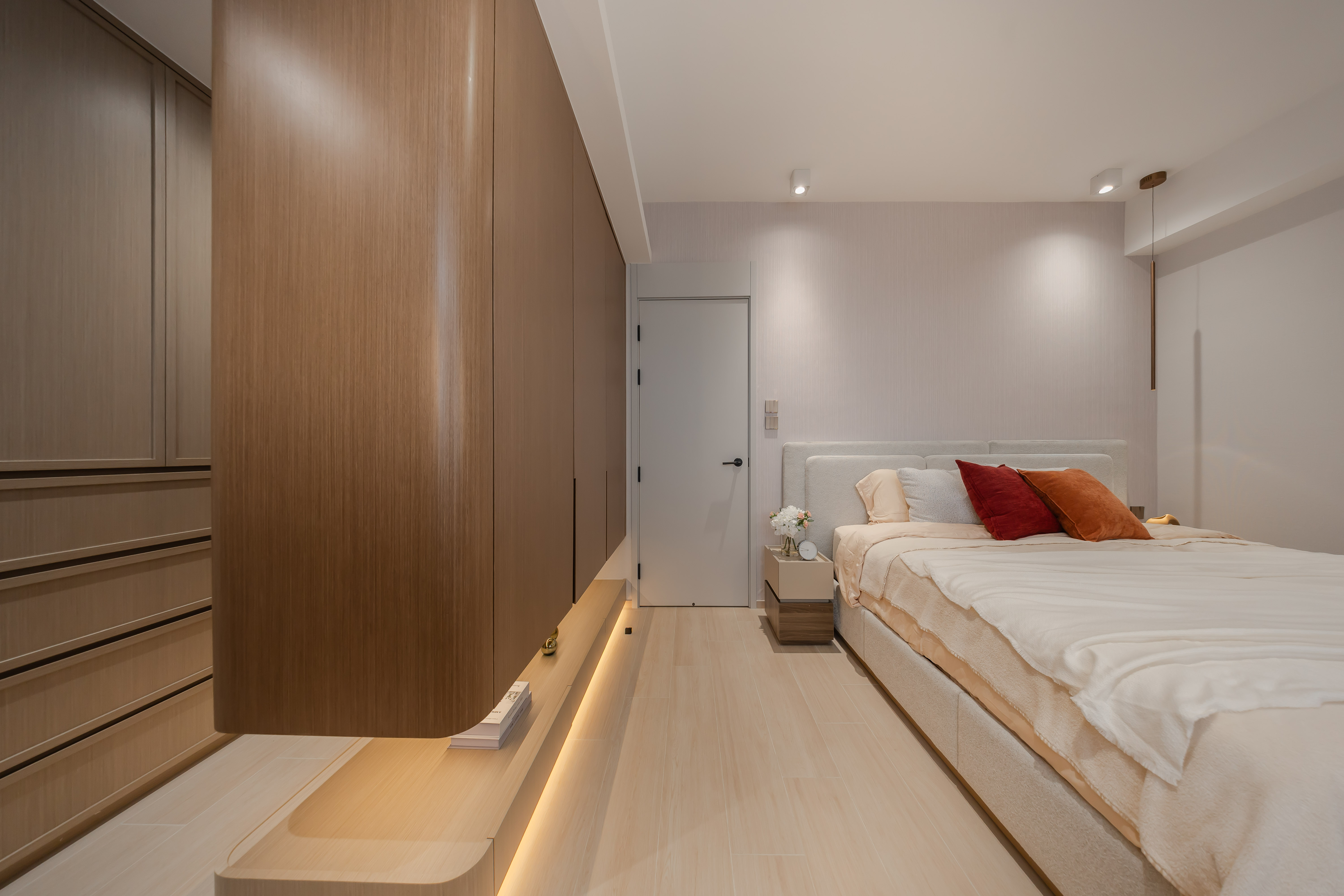 A modern bedroom featuring a large wooden wardrobe with curved edges, a bed with colorful pillows, and light wood flooring.