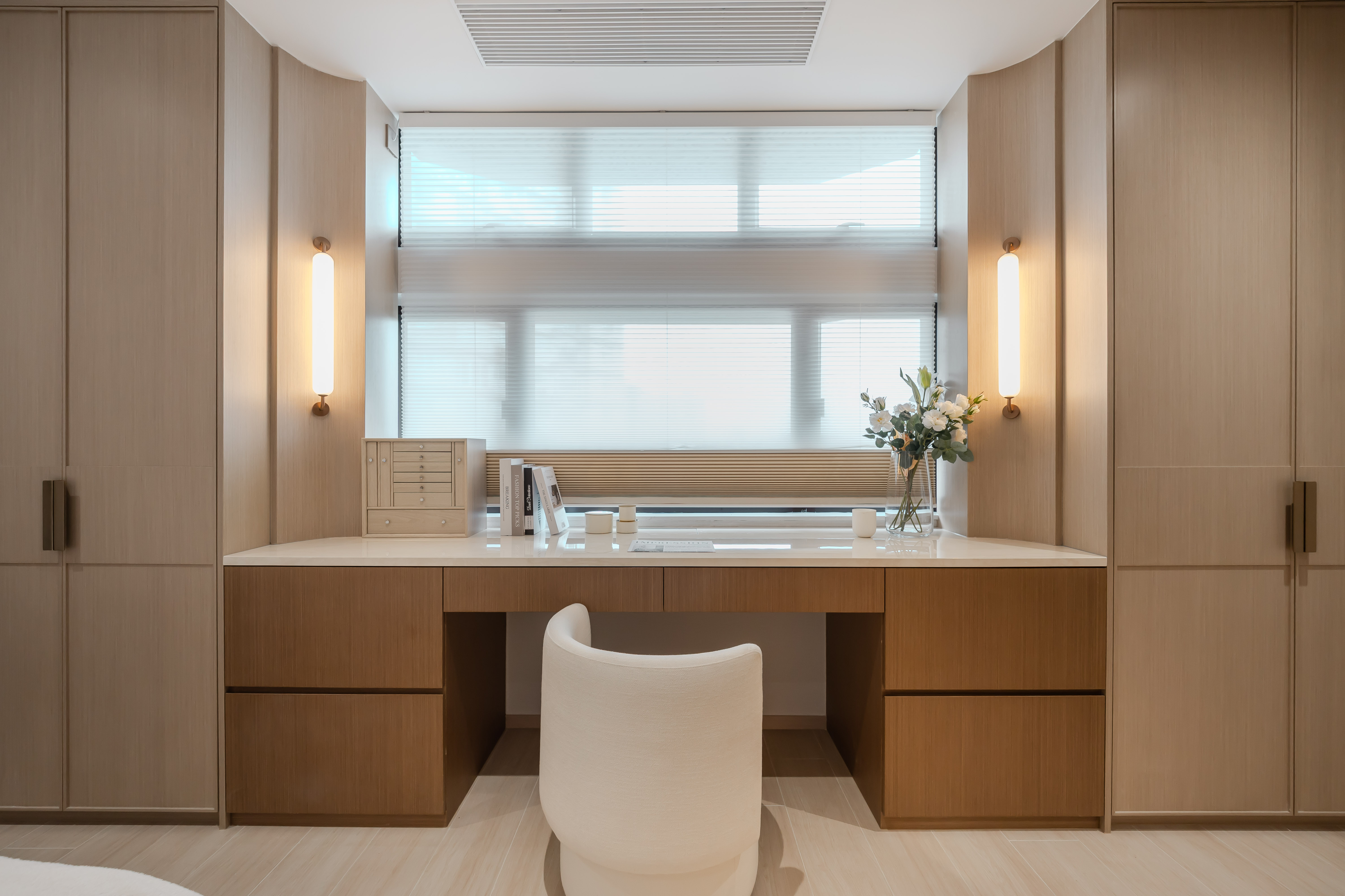 A bedroom window area featuring a wooden desk, tall wardrobes on both sides, a white chair, striped blinds, and wall lamps.