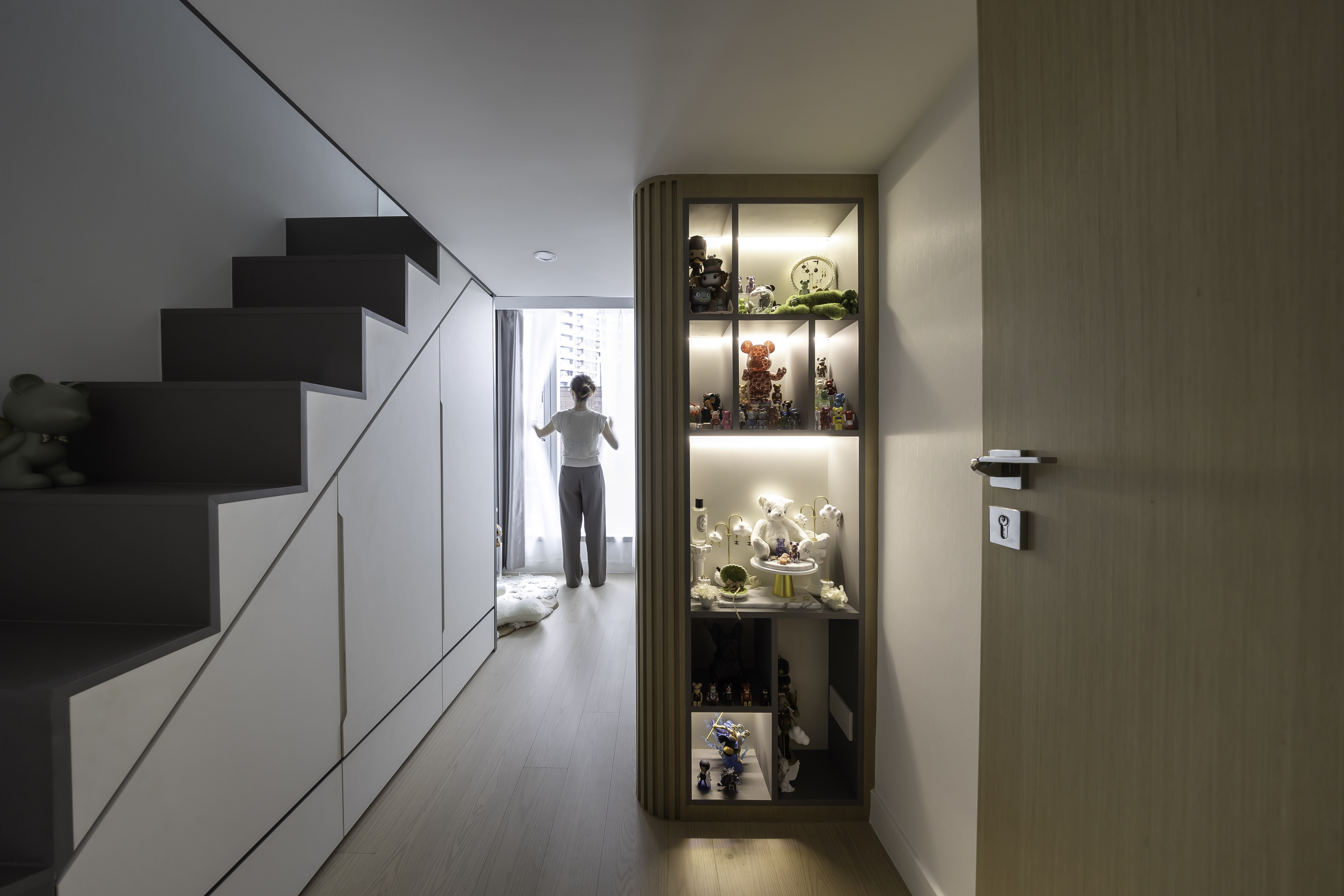 A hallway featuring stairs with built-in white storage cabinets underneath, and an illuminated glass display cabinet showcasing various figures.