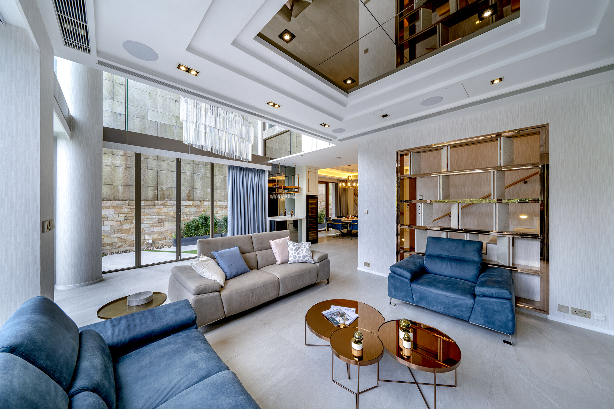 Spacious living room with high ceiling, mirrored ceiling panel, grey sofa, blue armchairs, and copper coffee tables.