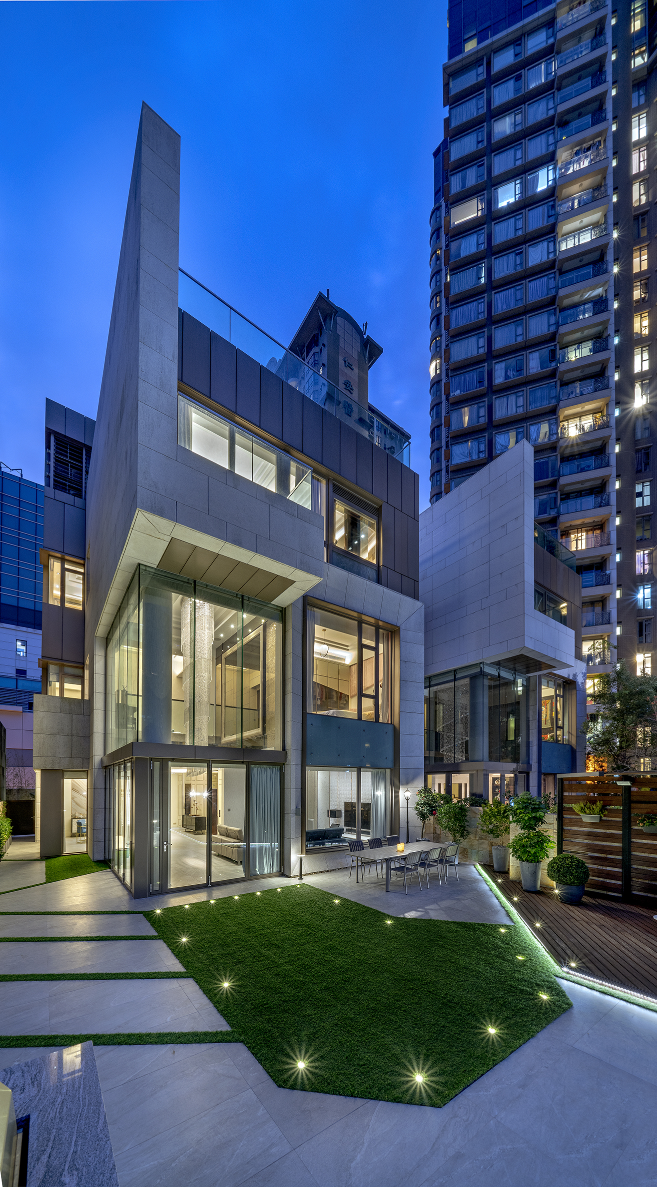 Exterior view of a modern multi-story house with large glass windows, featuring a courtyard with outdoor seating and artificial grass.