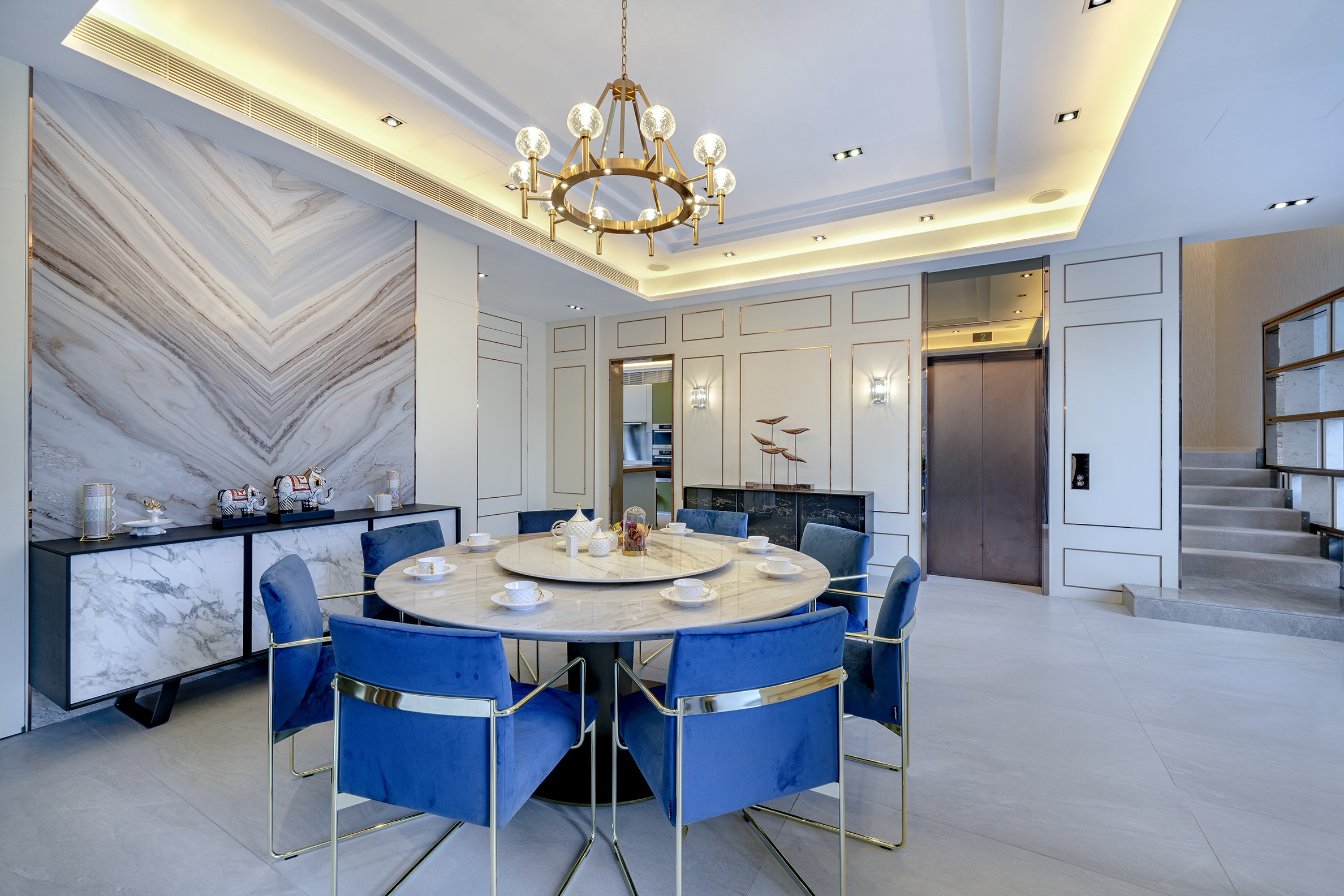 A dining room featuring a round marble table, blue velvet chairs, a gold chandelier, and a marble feature wall.