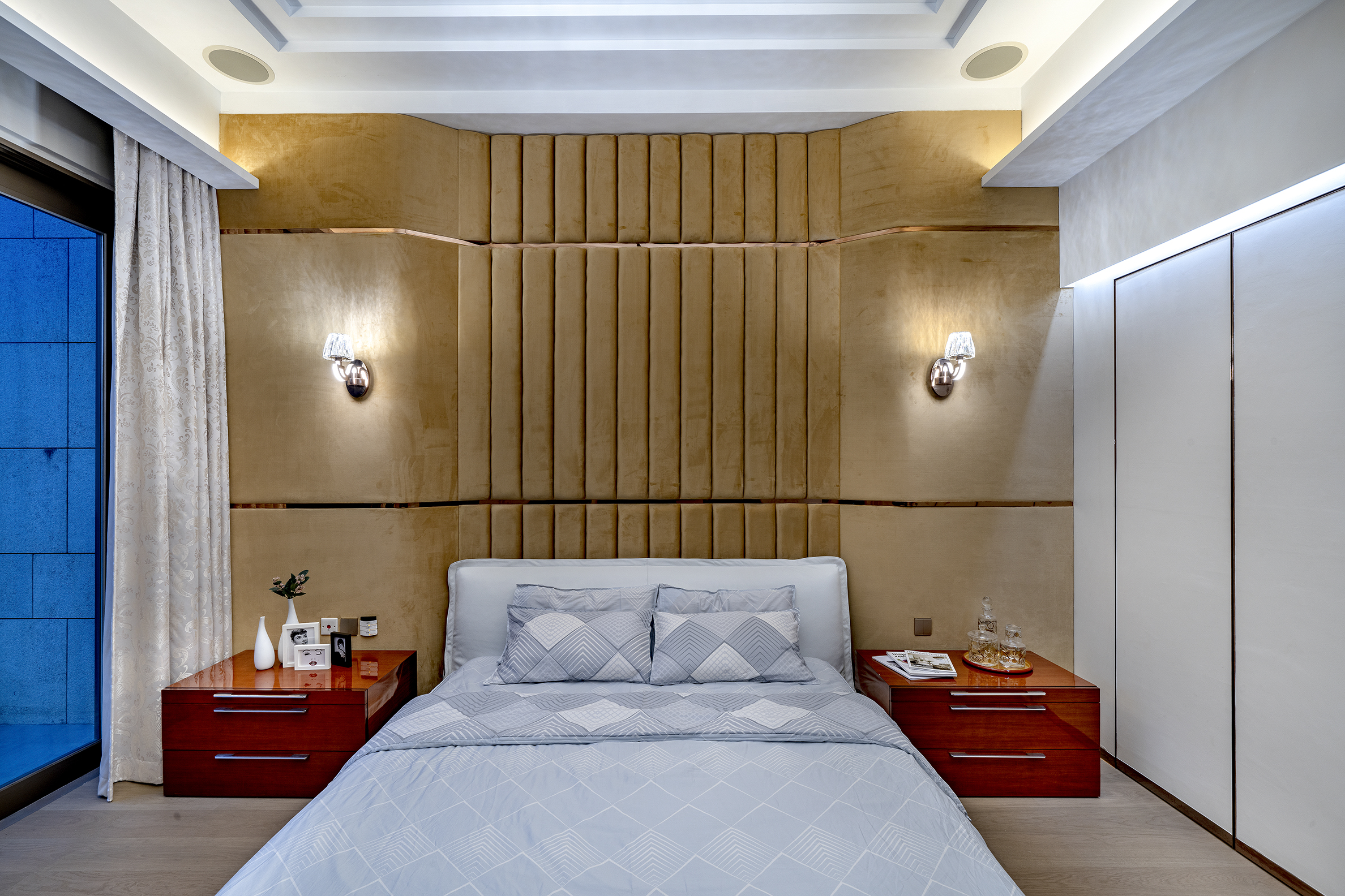 A bedroom featuring a large bed with grey patterned bedding, wooden nightstands, and a textured golden-brown upholstered headboard wall.