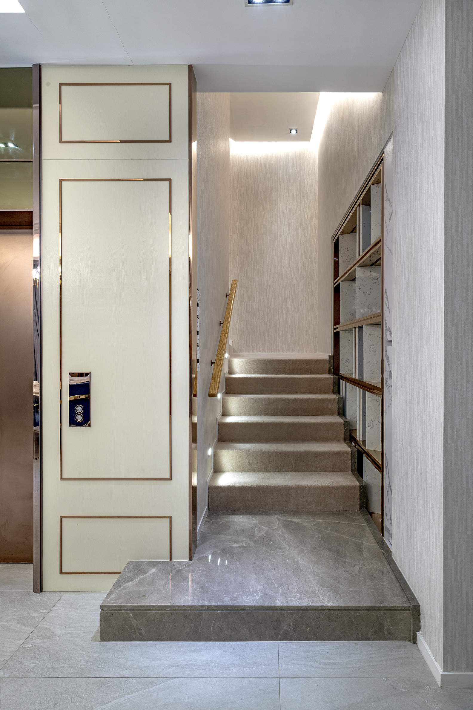 A modern staircase with carpeted steps, a wooden handrail, and a marble landing next to a white door with gold trim.