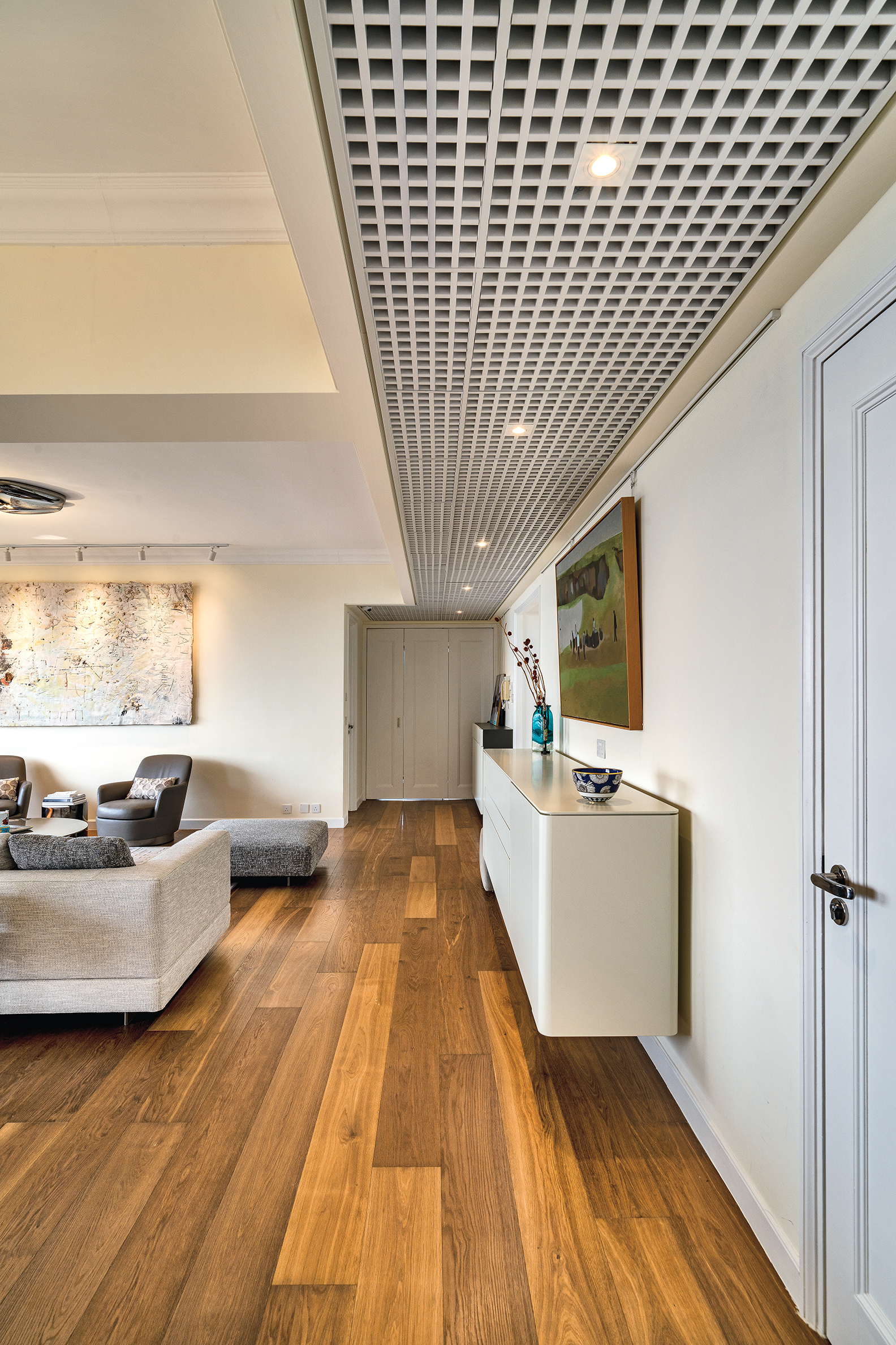 Living room and hallway with wooden flooring, a white grid ceiling panel, a floating white cabinet, and a grey sofa.