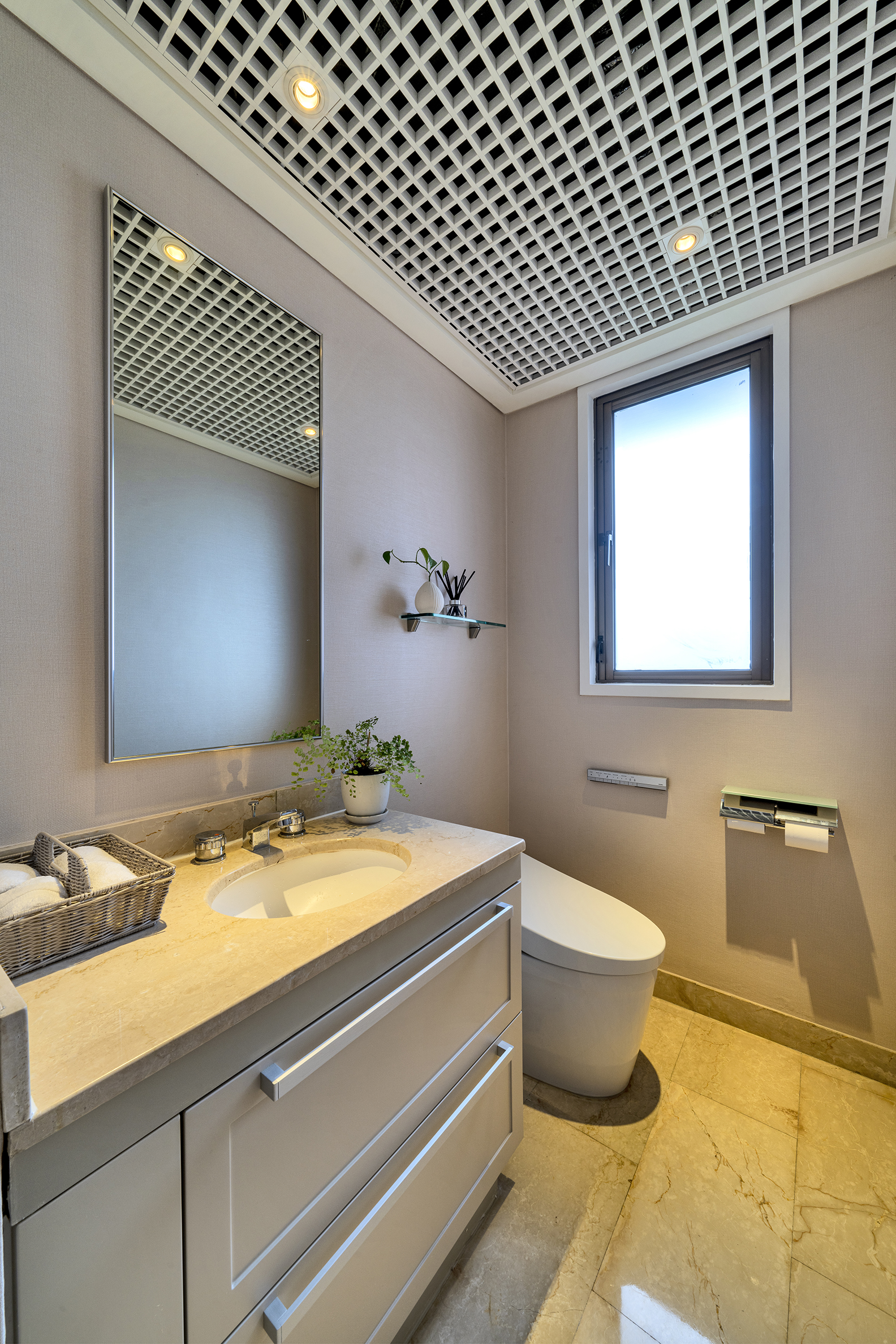 A modern bathroom featuring a beige stone vanity, a rectangular mirror, a white toilet, and a distinctive grid ceiling with recessed lights.