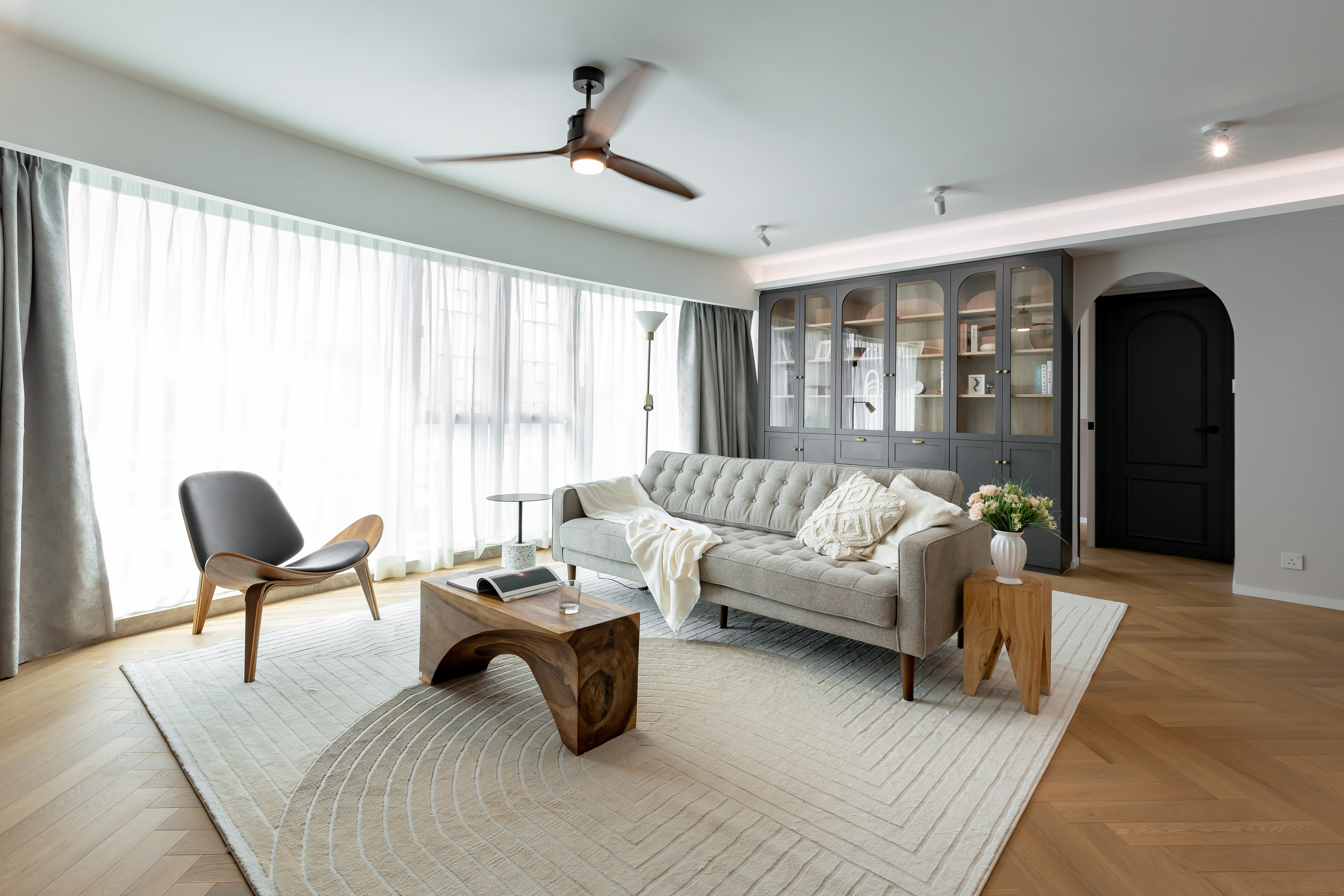 A bright living room with a grey tufted sofa, a wooden coffee table, a black lounge chair, and a large display cabinet.