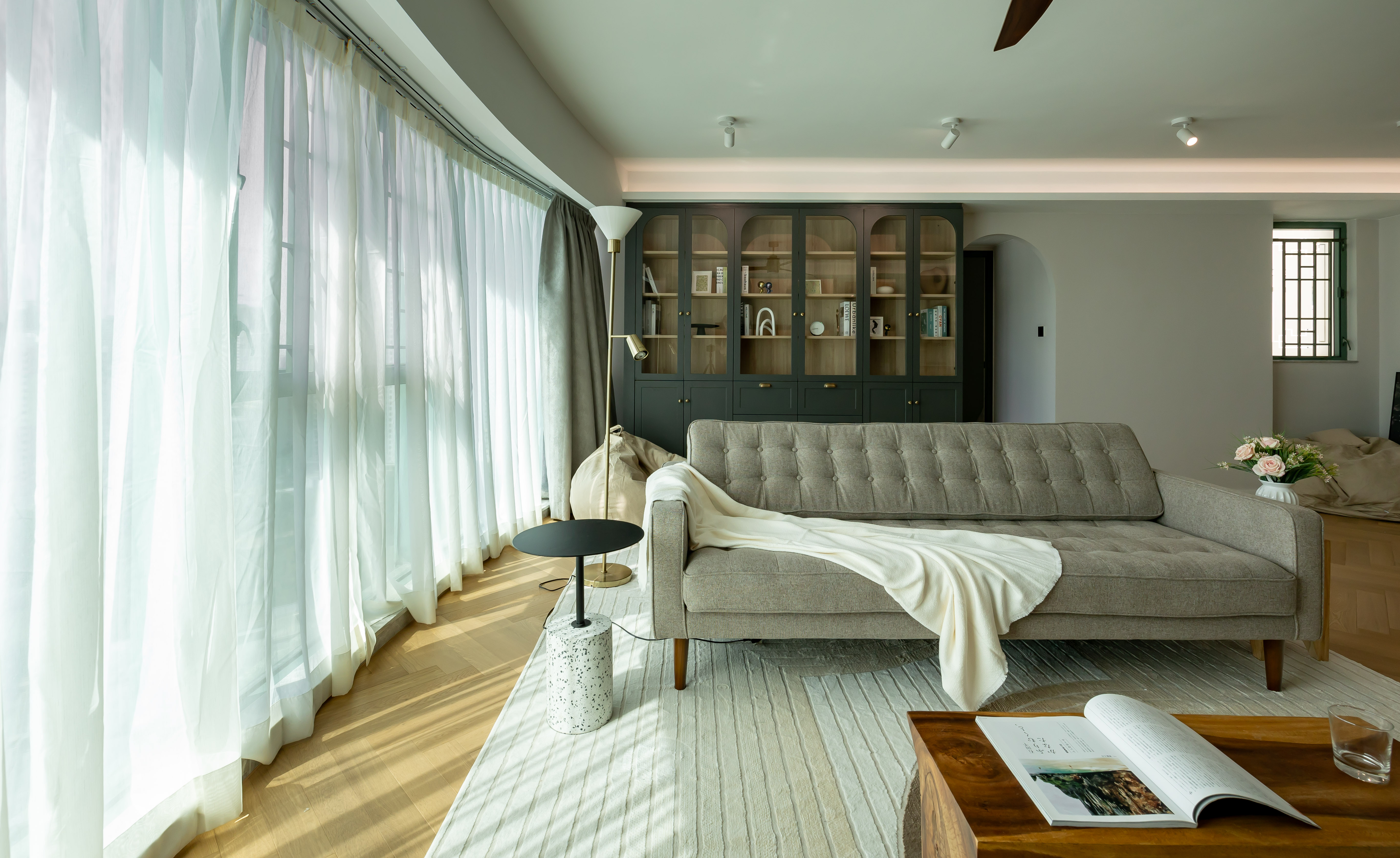 A bright living room with a grey tufted sofa, white curtains, and a dark green display cabinet.