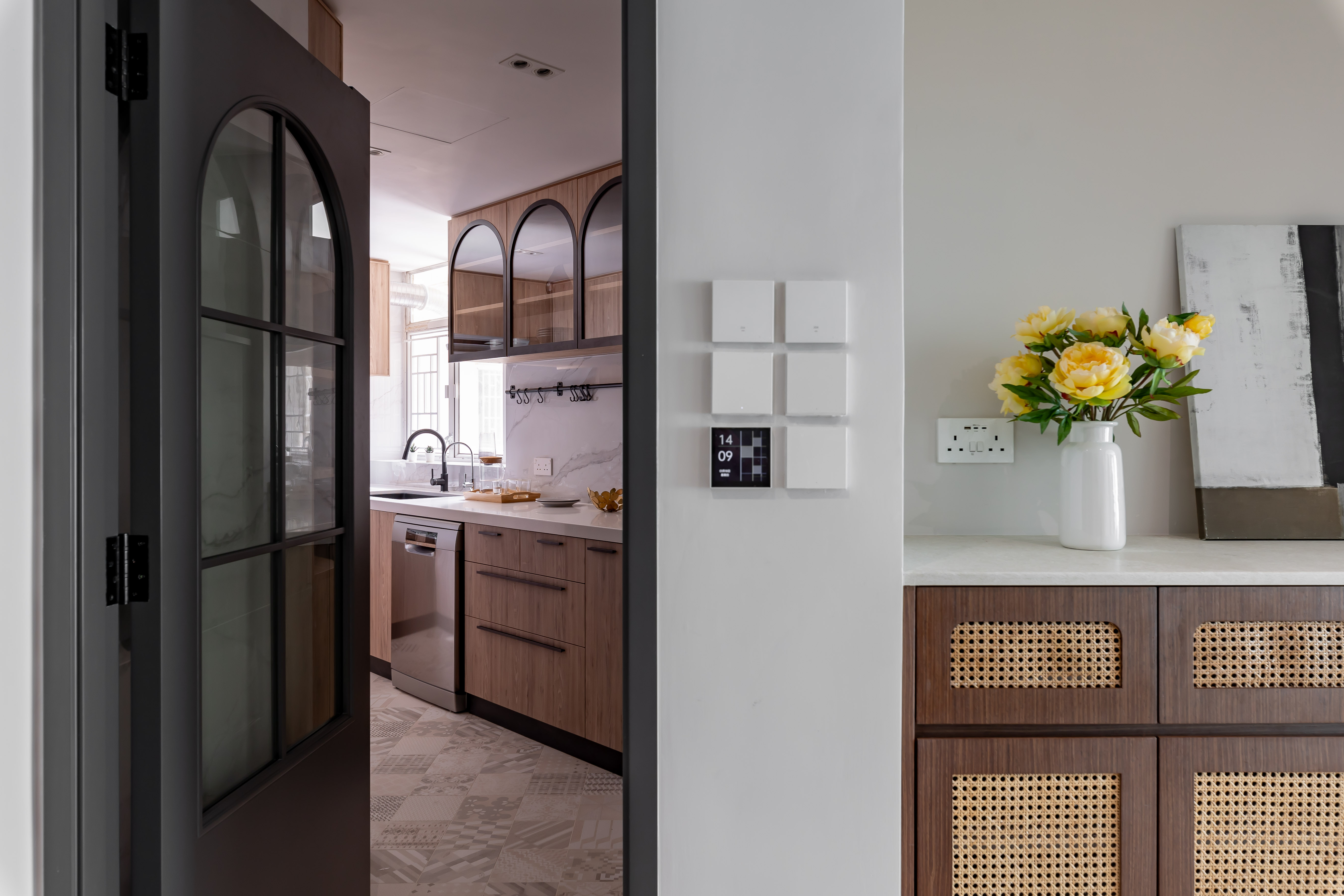 A view from a hallway looking into a kitchen with wooden cabinets, a black framed glass door, and a rattan cabinet.