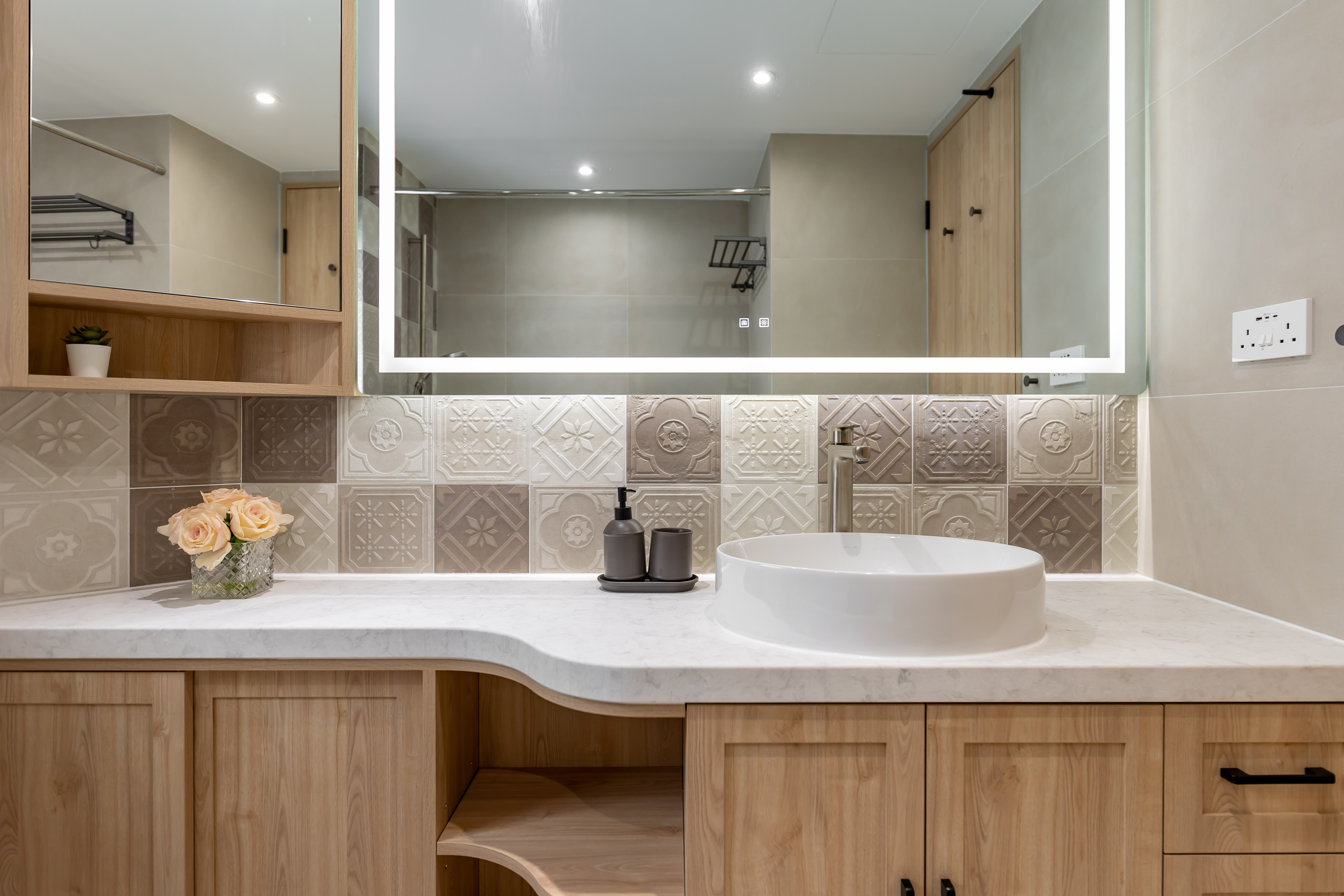 A modern bathroom vanity with a white vessel sink, wooden cabinets, patterned backsplash tiles, and a large illuminated mirror.