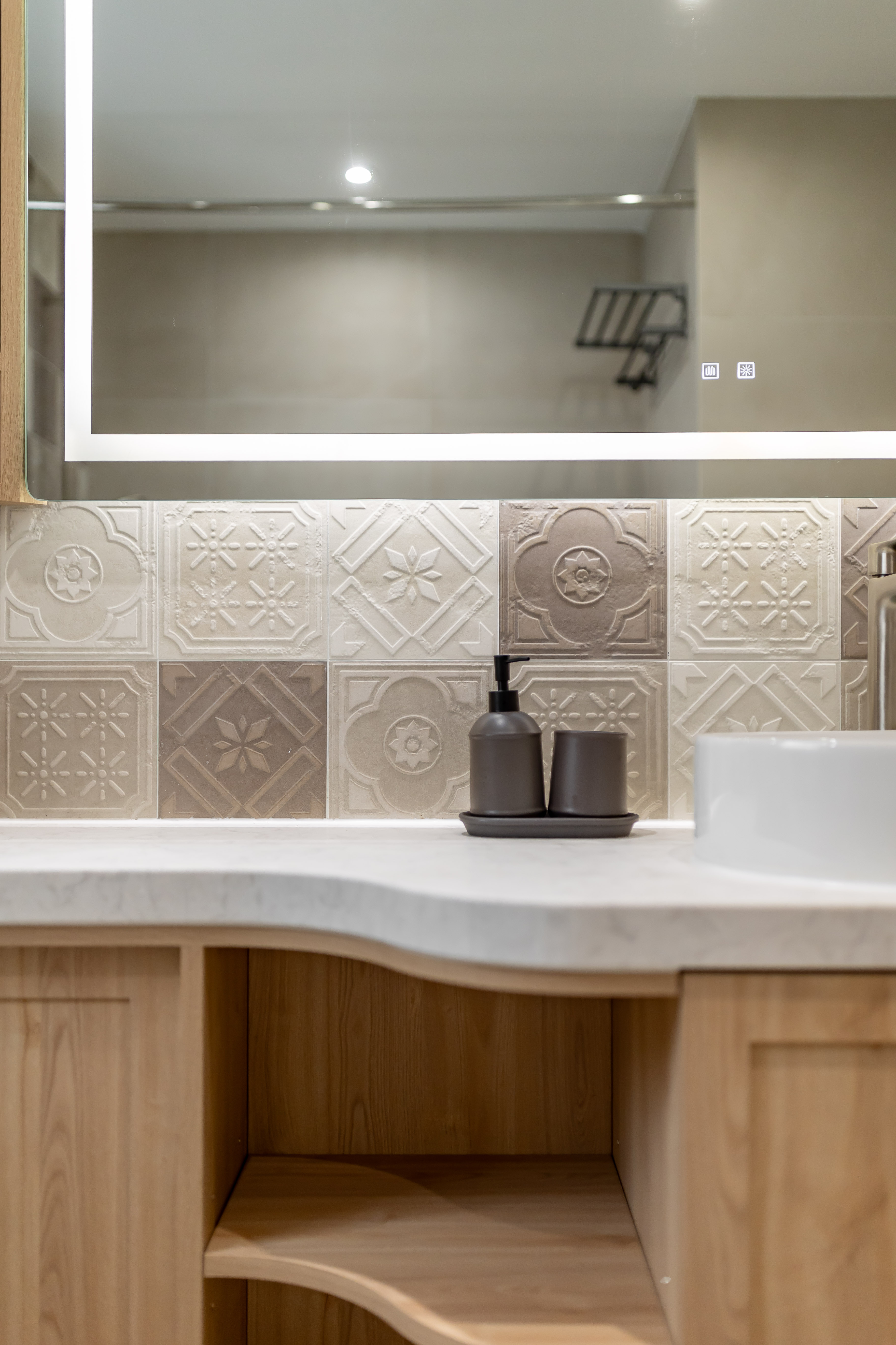 Bathroom featuring a lighted mirror cabinet, vintage patterned wall tiles, white stone countertop, wooden vanity, and grey soap dispenser.
