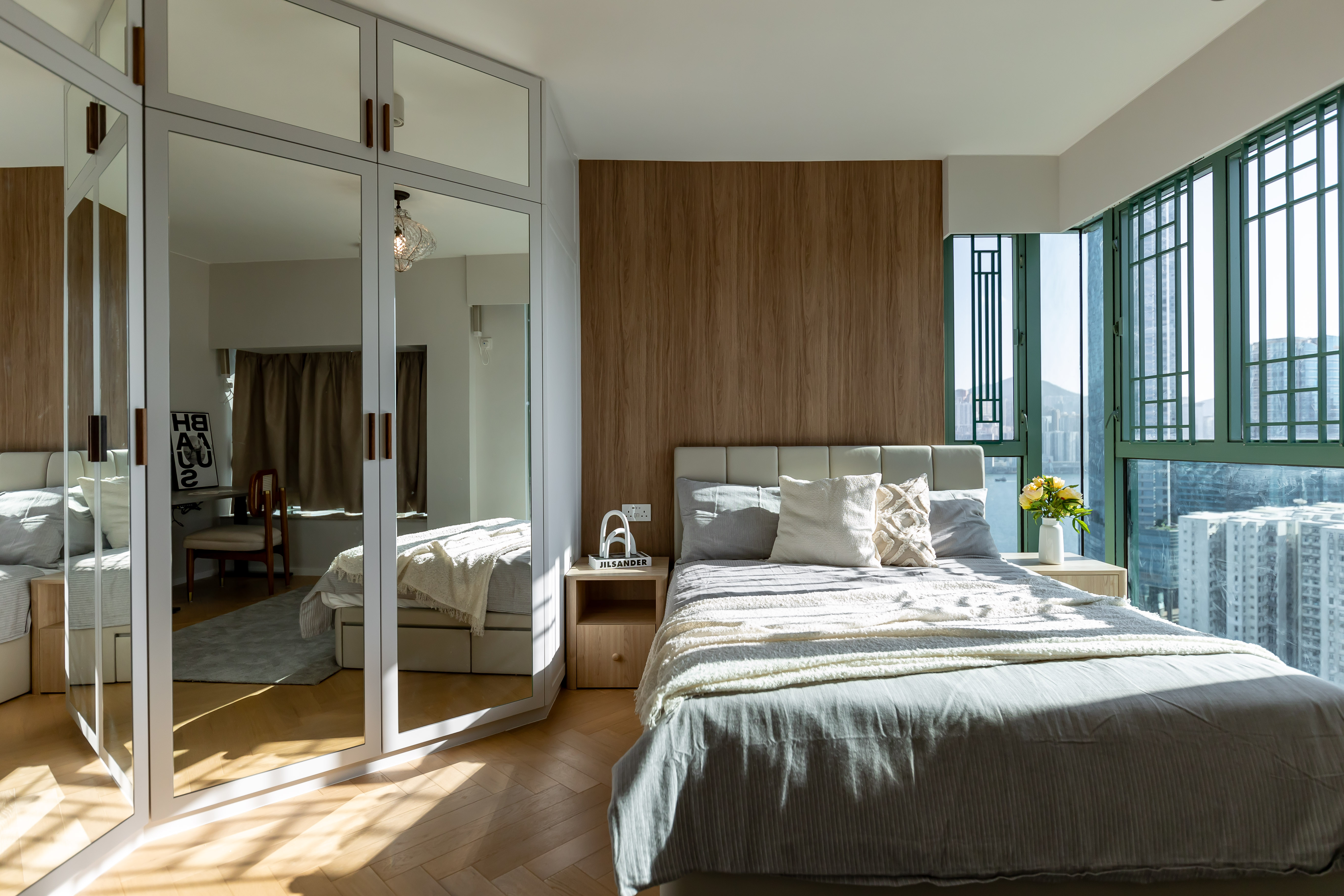 A bedroom featuring a large bed, wooden wall paneling, and a mirrored wardrobe reflecting the room and sunlight.