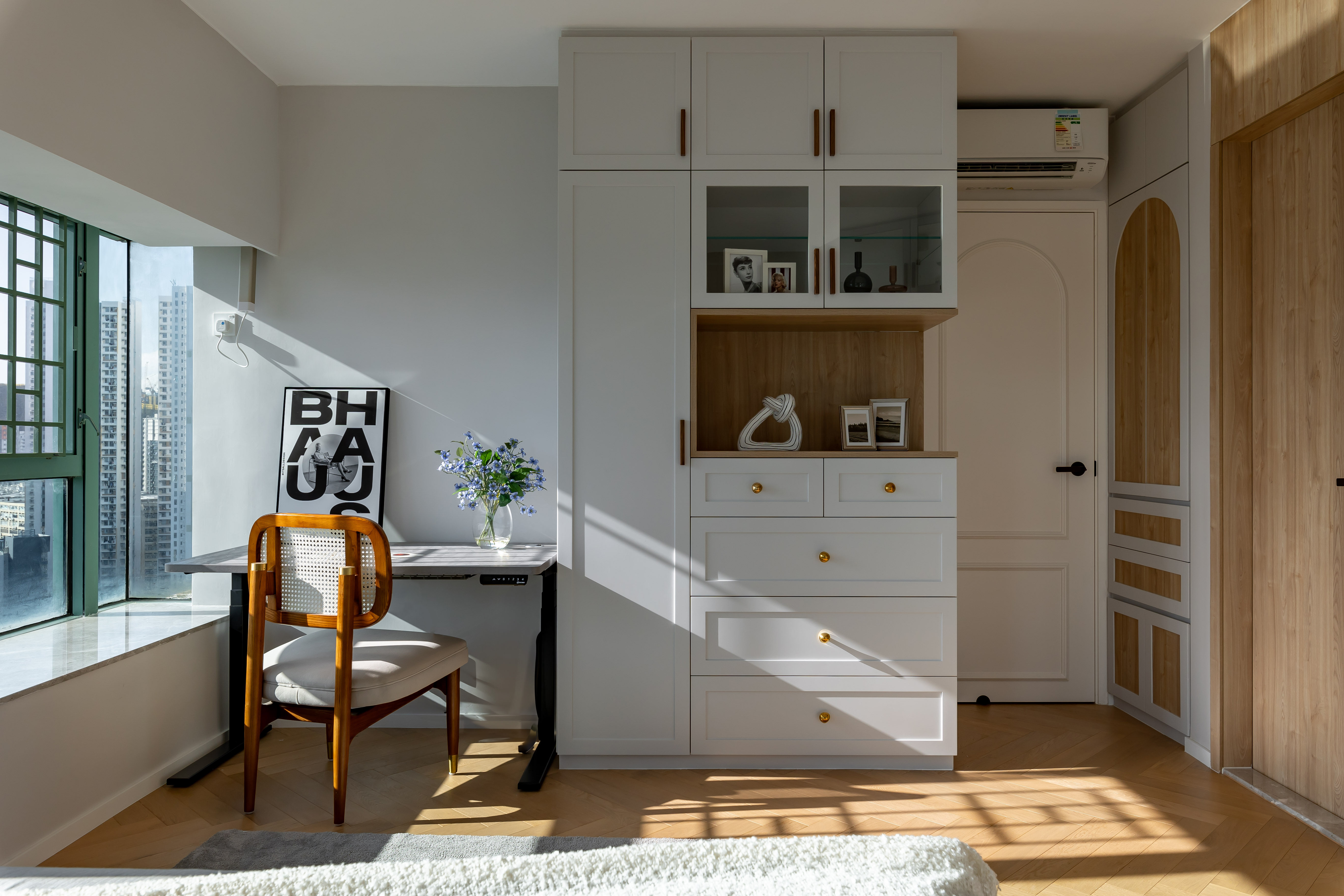 A bedroom featuring a white wardrobe, a desk by the window, and a wooden chair with sunlight on the wooden floor.