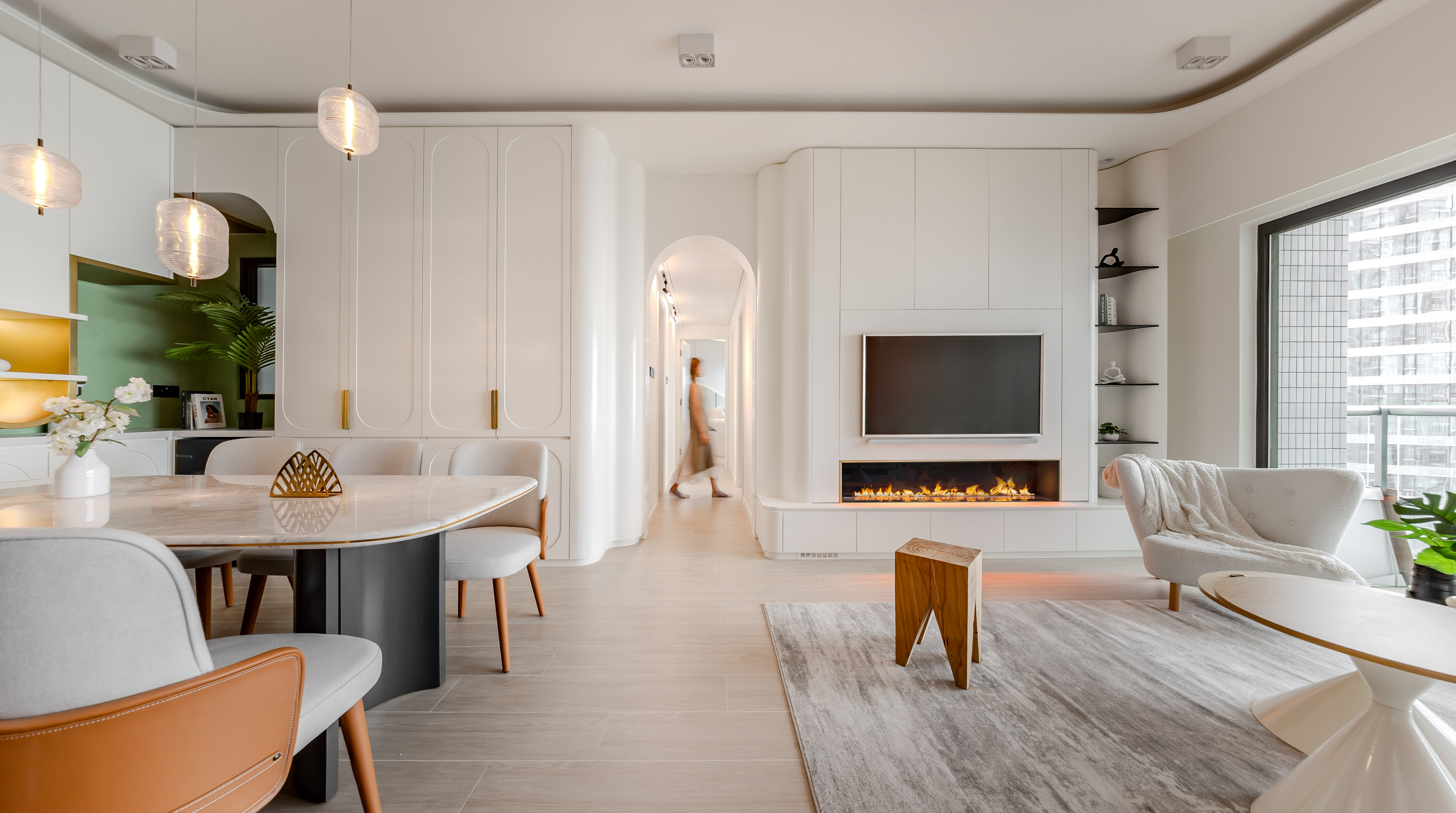 A bright living and dining room featuring white curved cabinets, a marble dining table, a wall-mounted TV, and a modern fireplace.