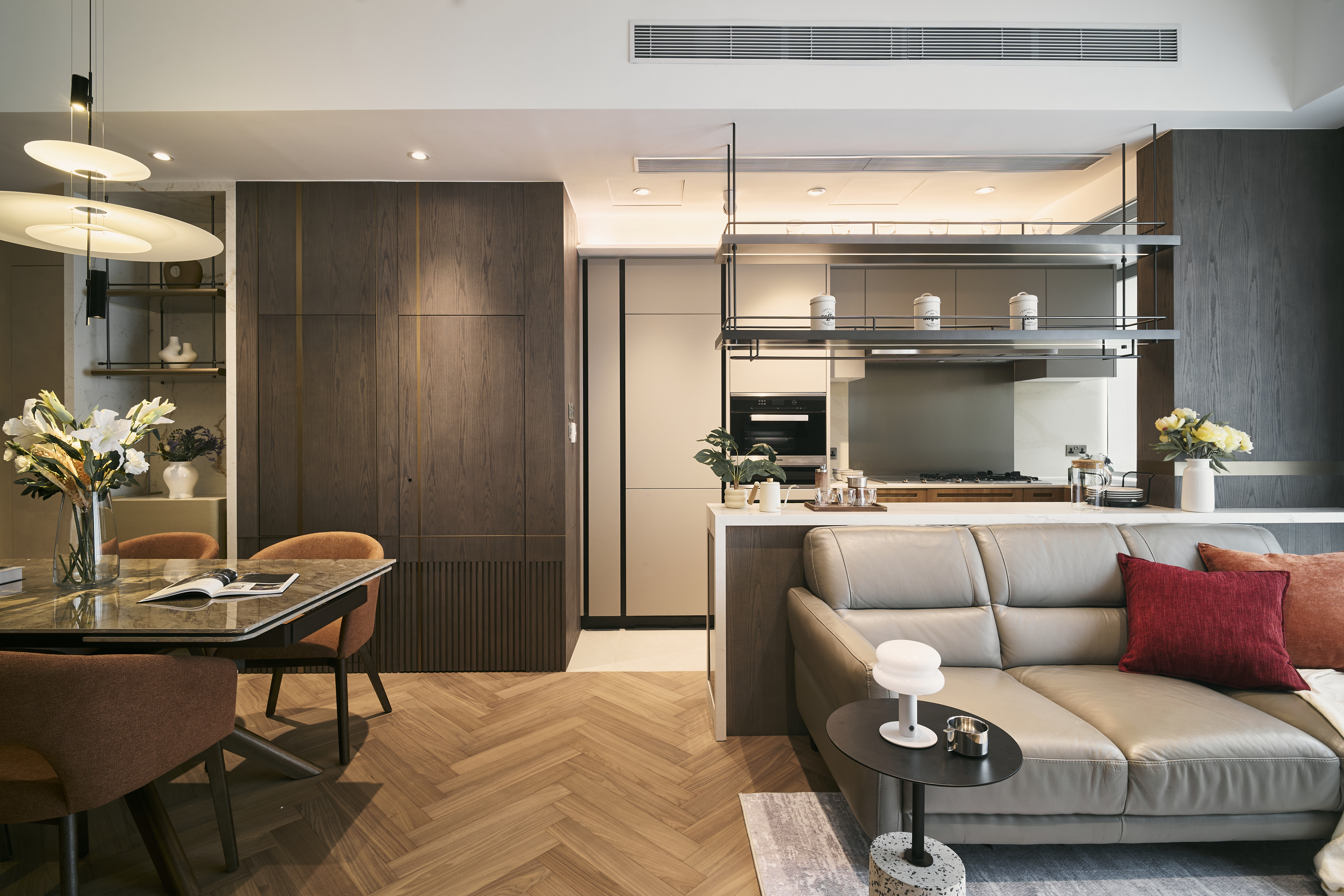 Modern living and dining room with open kitchen, featuring herringbone wood flooring, dark wood cabinets, and a grey leather sofa.